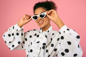 Studio Foto von afrikanisch schwarz Frau im stilvoll Kleid und Weiß Sonnenbrille. ziemlich lächelnd weiblich posieren Über Rosa Hintergrund.