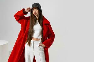 Mode Porträt von schön dünn Modell- im stilvoll Hut und rot Mantel posieren auf Weiß Hintergrund im Studio. tragen Ernte oben und Hose. foto