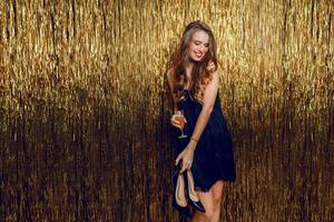 Neu Jahr feiern Party. einer Weiß glücklich verführerisch Frau posieren mit Glas von Champagner Über golden prickelnd Hintergrund. halten Paar von Schuhe im Hände. schwarz glamourös Kleid. foto
