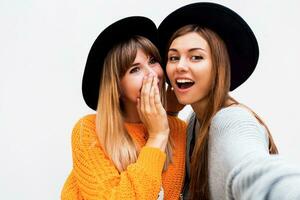 Freundschaft, Glück und Menschen Konzept. zwei lächelnd Mädchen Flüstern Klatsch auf Weiß Hintergrund. Orange Pullover, schwarz ähnlich Hüte. foto