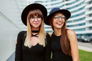 schließen oben Porträt von zwei modisch Frauen peinlich und Gehen zusammen draussen. tragen schwarz Outfit. Wunde Brille. Freundschaft Konzept. foto