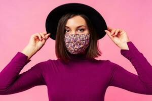 schön Frau tragen stilvoll schützend Gesicht Maske. Rosa Hintergrund. schwarz Hut. Studio Foto. foto