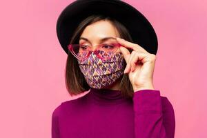 europäisch Modell- gekleidet schützend stilvoll Gesicht Maske. tragen schwarz Hut und Sonnenbrille. posieren Über Rosa Hintergrund im Studio. foto