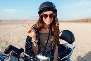 jung ziemlich heiter Frau Sitzung auf Motorrad auf das Strand und Show Zeichen, tragen stilvoll Ernte oben , Hemden, haben perfekt passen schlank gezähmt Körper und lange Haare. draussen Lebensstil Porträt. foto