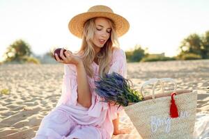 anmutig jung Frau mit blond wellig Haare im elegant Rosa Kleid genießen Picknick und Essen Früchte foto
