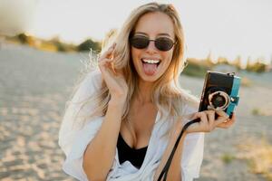 blond spielerisch Frau halten retro Kamera und haben Spaß auf sonnig Strand. Sonnenbrille, Weiß Outfit. foto