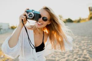 blond Frau halten retro Kamera und haben Spaß auf warm sonnig Strand. Sonnenbrille, Weiß Outfit. foto
