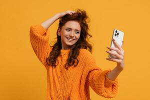 attraktiv rot Kopf Frau im stilvoll fallen Outfitm Sweatshirt und Jeans posieren Über Gelb Hintergrund im Studio. Herstellung selbst Porträt durch Smartphone. foto