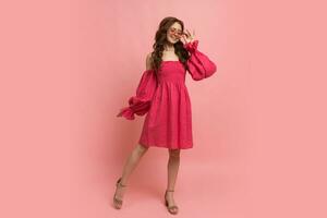 Mode Studio Porträt von stilvoll rothaarig Frau posieren im Rosa Pfandrecht Kleid mit Ärmel Über Rosa Hintergrund. voll Länge. foto