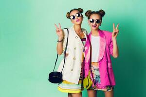 Mode Studio Bild von zwei jung Frauen im stilvoll beiläufig Frühling Outfit haben Spaß, Show Zunge. hell modisch Farben, stilvoll Frisur mit Gebäck , cool Sonnenbrille. freunde Porträt. foto