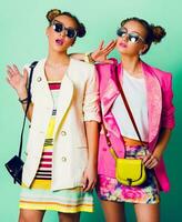Mode Studio Bild von zwei jung Frauen im stilvoll beiläufig Frühling Outfit haben Spaß, Show Zunge. hell modisch Pastell- Farben, stilvoll Frisur mit Gebäck , cool Sonnenbrille. freunde Porträt. foto