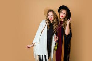 zwei Modelle mit blond wellig Frisur im wolle und Stroh Hut tragen gestreift Poncho. amerikanisch oder australisch Hippie Bohemien Stil. Mädchen Lachen und genießen Zeit .Beige Hintergrund. foto