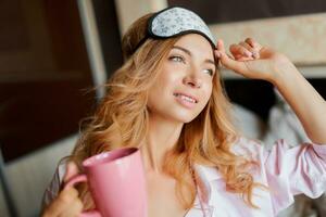 ziemlich Frau mit offen Lächeln posieren im Auge Maske beim Zuhause und Trinken heiß Tee. gemütlich Zuhause Atmosphäre. foto