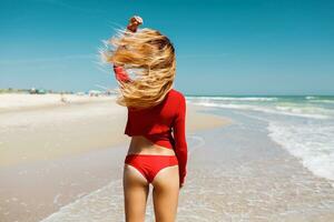 schließen oben draussen Schuss von jung sexy blond Frau im rot Bikini, Sonnenbaden beim Meer Ufer. Ozean Strand Hintergrund. perfekt schlank Körper und Hintern mit Sand. Ferien und Reise Konzept. Foto von zurück.