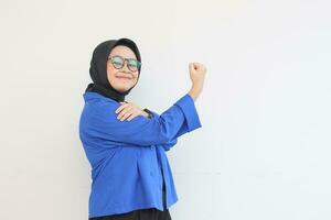 schön jung asiatisch Muslim Frau, tragen Brille und Blau Blazer mit stark Geste durch Heben ihr Waffen und Muskeln foto