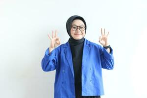 schön jung asiatisch Muslim Frau, tragen Brille und Blau Blazer mit Hand Geste von die Genehmigung oder in Ordnung foto