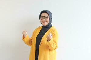 schön jung asiatisch Muslim Frau, tragen Brille und Gelb Blazer mit glücklich Ausdruck während Zusammenpressen Faust foto