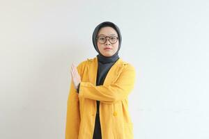 schön jung asiatisch Muslim Frau, tragen Brille und Gelb Blazer zeigen halt Geste während Gerade Gesicht Ausdruck foto