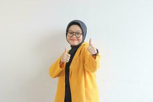 schön jung asiatisch Muslim Frau, tragen Brille und Gelb Blazer mit Hand Geste von die Genehmigung oder in Ordnung foto