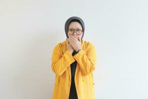 schön jung asiatisch Muslim Frau, tragen Brille und Gelb Blazer mit schockiert Ausdruck während Abdeckung Mund foto