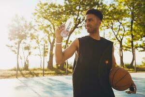 Nahansicht Porträt von jung schwarz Mann tun Sport im Morgen, Trinken Wasser auf Basketball Gericht foto