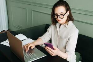 produktiv Gespräche. Frau balancieren Laptop und Smartphone im Video Forderung. Multitasking Freude. beschäftigt Frau im Brille jongliert Video Anruf auf Laptop und Smartphone mit ein Lächeln foto