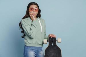 Überraschtes Teenager-Mädchen mit Sonnenbrille, das ein Skateboard hält foto