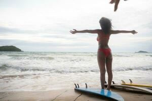 zurück Aussicht von jung Frau Stehen auf das Strand mit ihr Surfbrett foto