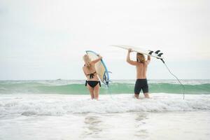 jung Mann und Frau halten Surfbretter auf ihr Köpfe und gehen in das Meer zu Surfen foto