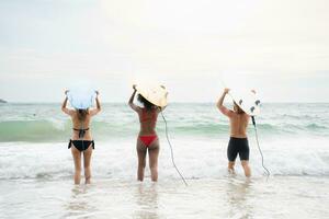 Rückseite Aussicht von zwei Frauen und jung Mann halten Surfbretter auf ihr Köpfe und gehen in das Meer zu Surfen foto