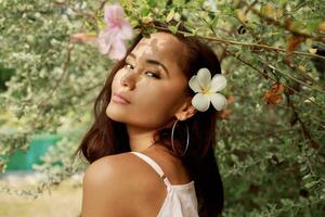 anmutig asiatisch Frau mit perfekt Haut und Hibiskus Blume im Haare posieren Über Holz Mauer und tropisch Pflanzen. Sommer- Mode Porträt. foto