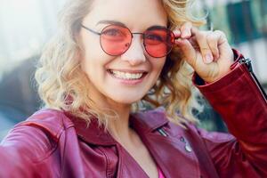 glücklich blond Frau posieren auf modern Straßen, Trinken Kaffee oder Cappuccino. stilvoll Herbst Outfit, Leder Jacke und gestrickt Pullover. städtisch Hintergrund. Rosa Sonnenbrille. foto
