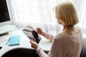 lächelnd reifen Frau mit Handy, Mobiltelefon Telefon während Arbeiten durch Computer beim heim. foto