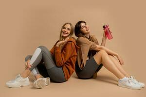 zwei Lachen sportlich Frauen chillen nach Ausbildung, Sitzung auf Fußboden im Studio auf Beige Hintergrund. foto