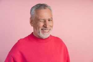 neugieriger, älterer Mann in einem hellen Pullover zwinkert auf rosa Hintergrund foto