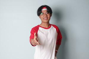 Porträt von attraktiv asiatisch Mann im T-Shirt mit rot und Weiß Band auf Kopf, erreichen aus zum ein Handschlag und grüßen jemand. isoliert Bild auf grau Hintergrund foto