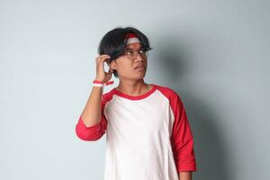 Porträt von attraktiv asiatisch Mann im T-Shirt mit rot und Weiß Band auf Kopf, Stehen gegen grau Hintergrund, Denken Über Frage mit Hand auf Kinn foto