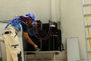 afrikanisch amerikanisch Arbeiter fegen Weg lose Trümmer angesammelt um Luft Conditioner Base. trainiert Ingenieur Reinigung hvac System Körper und Steuerung Panel mit Abstauben Bürste Werkzeug zu entfernen lose Schmutz foto