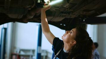 Frau Arbeiten auf suspendiert Auto im Garage, Überprüfung Teile während jährlich Untersuchung. Fachmann im Reparatur Geschäft Arbeitsplatz Gehen unterhalb Fahrzeug, Scannen es mit Arbeit Licht foto