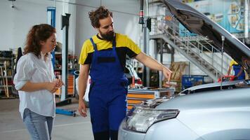 Dolly aus Schuss von Handwerker assistieren Klient mit Auto Instandhaltung im Reparatur Geschäft. Mitarbeiter im Garage Arbeitsplatz suchen Über Automobil Teile mit Frau, reparieren ihr Fahrzeug während jährlich Untersuchung foto