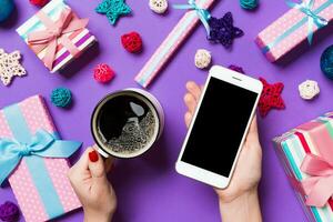 Draufsicht einer Frau, die ein Telefon in der einen Hand und eine Tasse Kaffee in der anderen Hand auf violettem Hintergrund hält. Weihnachtsschmuck und Spielzeug. neujahrsferienkonzept. Attrappe, Lehrmodell, Simulation foto
