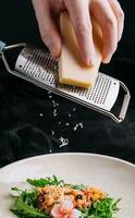 das Prozess von Kochen warm Salat. Hand von das Koch reibt das Käse auf ein Reibe. foto