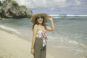 ein glücklich jung asiatisch Frau tragen Strand Hut ist posieren zu das Kamera beim das Strand im gunungkidul, Indonesien foto