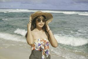 ein glücklich jung asiatisch Frau tragen Strand Hut ist posieren zu das Kamera beim das Strand im gunungkidul, Indonesien foto