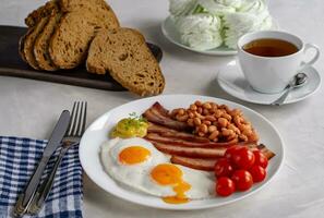 herzhaft Frühstück mit durcheinander Eier, Speck, Bohnen, Kirsche Tomaten, hausgemacht brot, Tee und Marshmallows auf ein Beton Arbeitsplatte. Englisch Frühstück Konzept. foto