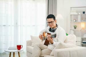 asiatisch Mann tragen Brille und Kopfhörer spielen Gitarre während Sitzung auf Sofa im das Leben Zimmer beim heim. asiatisch Mann Schreiben Lied während spielen Gitarre beim heim. komponieren Lied Musik- Konzept. foto