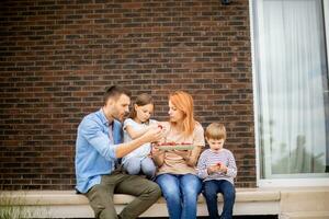 Familie mit ein Mutter, Vater, Sohn und Tochter Sitzung draußen auf Schritte von ein Vorderseite Veranda von ein Backstein Haus und Essen Erdbeeren foto