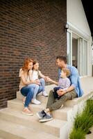 Familie mit ein Mutter, Vater, Sohn und Tochter Sitzung draußen auf das Schritte von ein Vorderseite Veranda von ein Backstein Haus foto