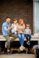 Familie mit ein Mutter, Vater, Sohn und Tochter Sitzung draußen auf Schritte von ein Vorderseite Veranda von ein Backstein Haus und Essen Erdbeeren foto