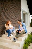 Familie mit ein Mutter, Vater, Sohn und Tochter Sitzung draußen auf das Schritte von ein Vorderseite Veranda von ein Backstein Haus foto